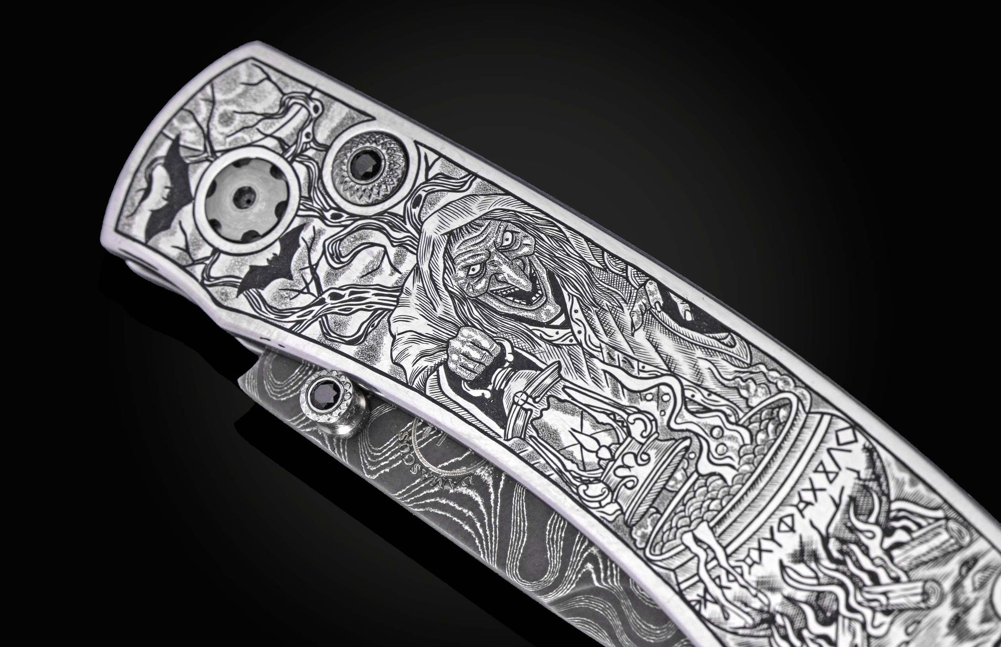 Silver folding knife with intricate Halloween-themed engraving featuring a witch, bats, and occult symbols, side view on black background