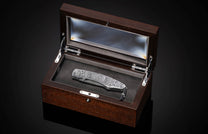 Luxury folding knife with intricate Halloween-themed engraving in silver, displayed in a dark wood presentation box with glass lid and black leather lining.