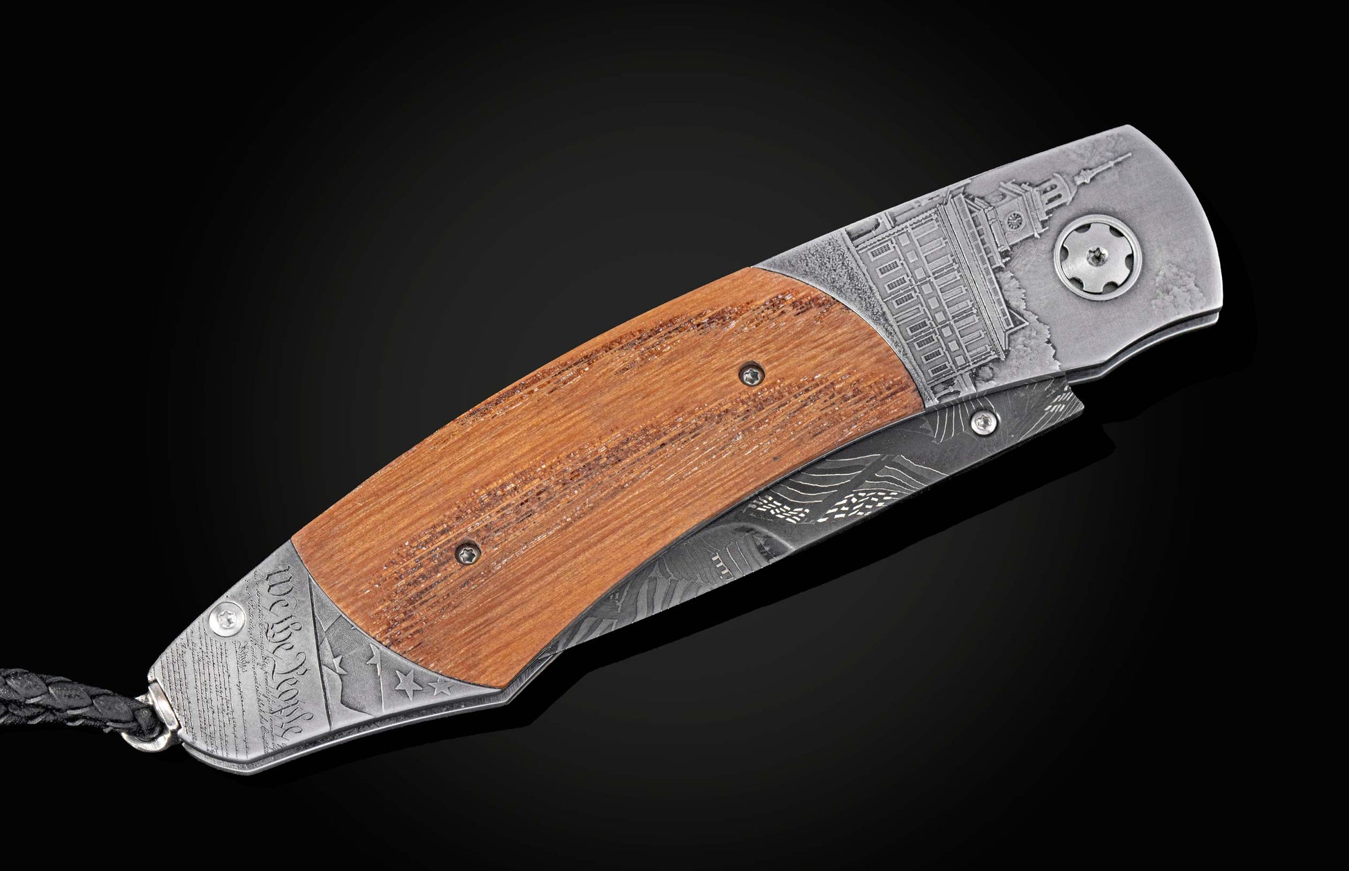 Closed folding knife with a brown wood handle, engraved metal featuring Independence Hall, 'We The People' script, and a patterned blade with a braided lanyard.