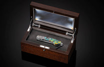 Luxury folding knife with vibrant mosaic handle and engraved blade, displayed in dark wood presentation box with glass lid, top-down view