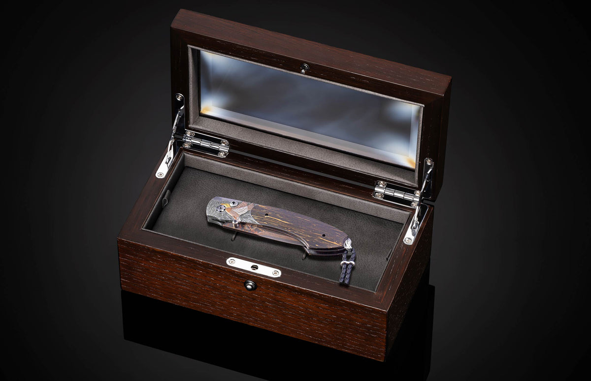 Folding knife with an engraved eagle on a dark wood handle and braided lanyard, displayed in an open dark brown display box with black interior.