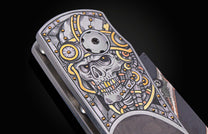 Silver engraved folding knife handle with an intricate steampunk-style skull design featuring gold and copper accents, seen close-up.