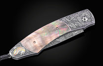 Luxury folding knife with mother-of-pearl inlay and engraved horse motifs, by B12, featuring damascus steel blade and detailed silver hardware, side view on black background