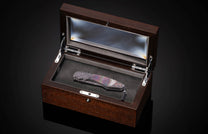 Luxury folding knife with iridescent mother-of-pearl inlay and engraved metal handle, by B12, displayed in dark wood presentation box with glass lid, top-down view