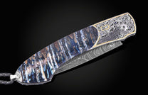 Luxury folding knife with blue marbled handle and damascus blade, featuring engraved raven motif and gemstone accents, side view on black background
