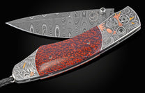 A folding knife with a wavy Damascus blade and textured red handle, featuring silver engravings of a skull, wings, and motorcycle with gold and copper accents.