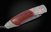 Custom folding knife with reddish-brown textured handle, engraved silver bolster featuring a skull, flames, and feathers, and a Damascus blade.