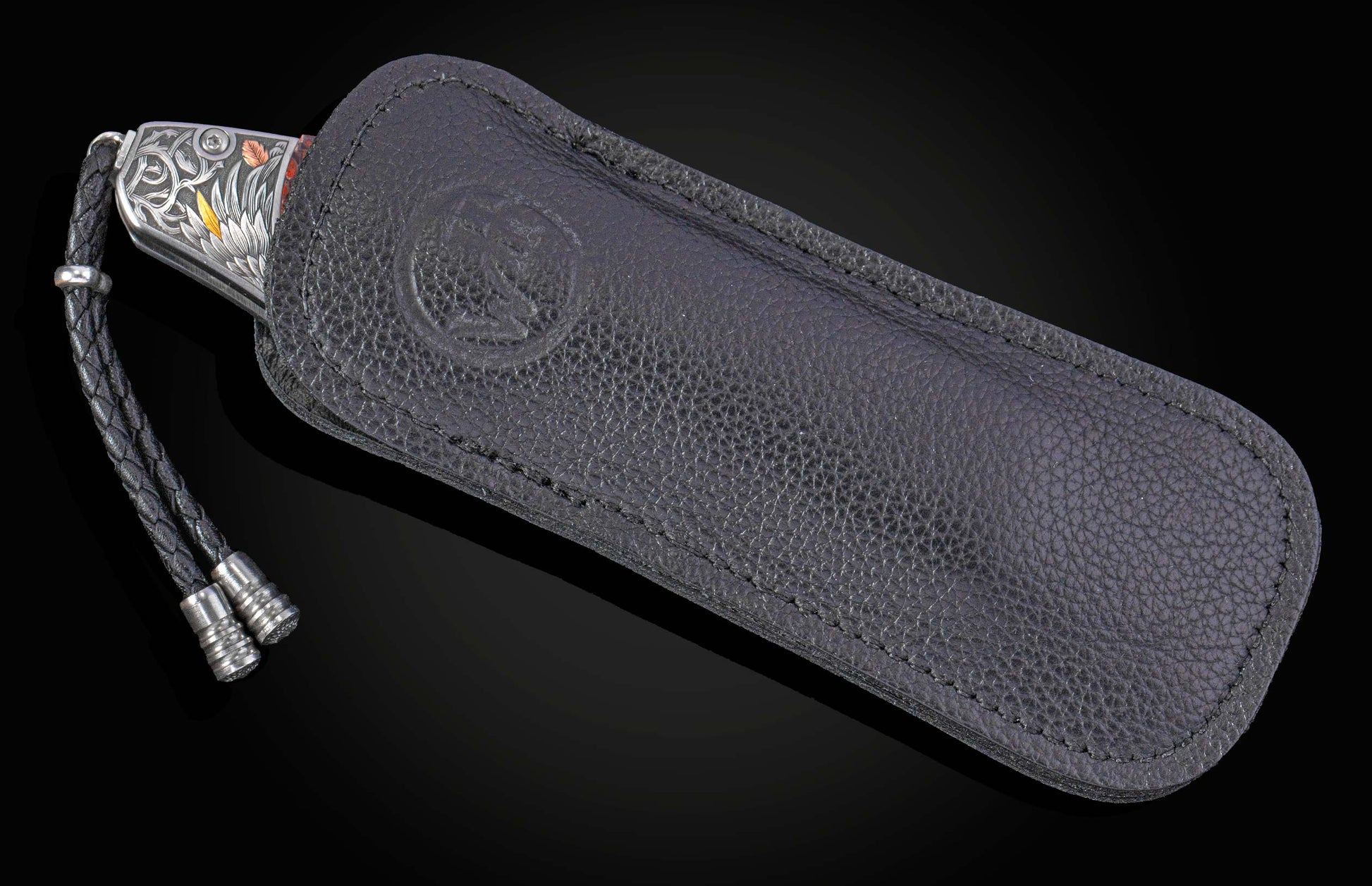 Silver-toned folding knife with intricate floral engraving and yellow/copper accents, and a black braided lanyard, partially in a black pebbled leather sheath with an embossed logo.