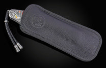 Silver-toned folding knife with intricate floral engraving and yellow/copper accents, and a black braided lanyard, partially in a black pebbled leather sheath with an embossed logo.