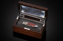 Folding knife with red textured handle, engraved silver metal, and a patterned blade, displayed in an open dark brown wooden box with a black padded interior.