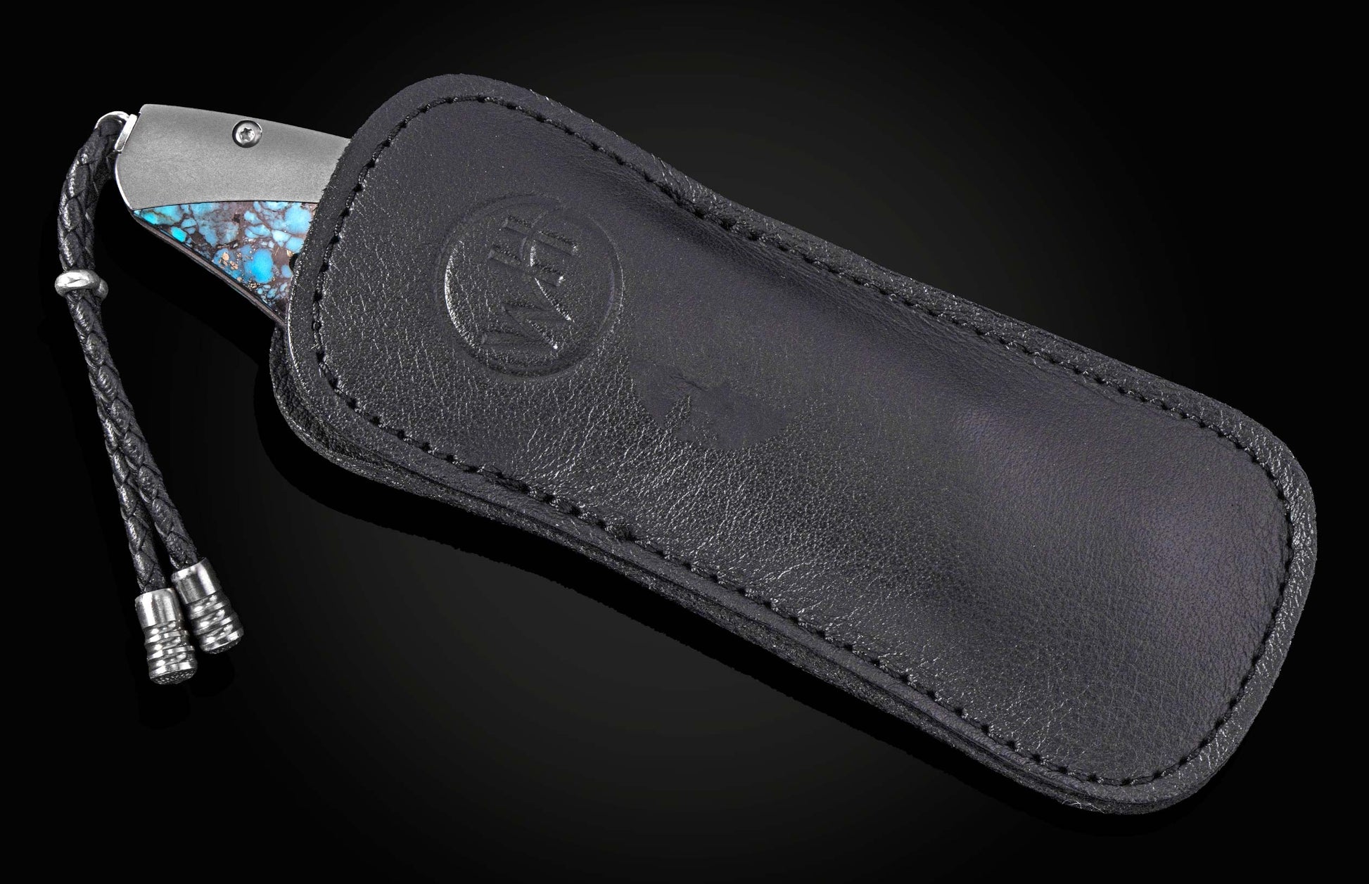 Luxury folding knife with turquoise inlay handle by B12, partially sheathed in black leather case with embossed logo and braided cord.