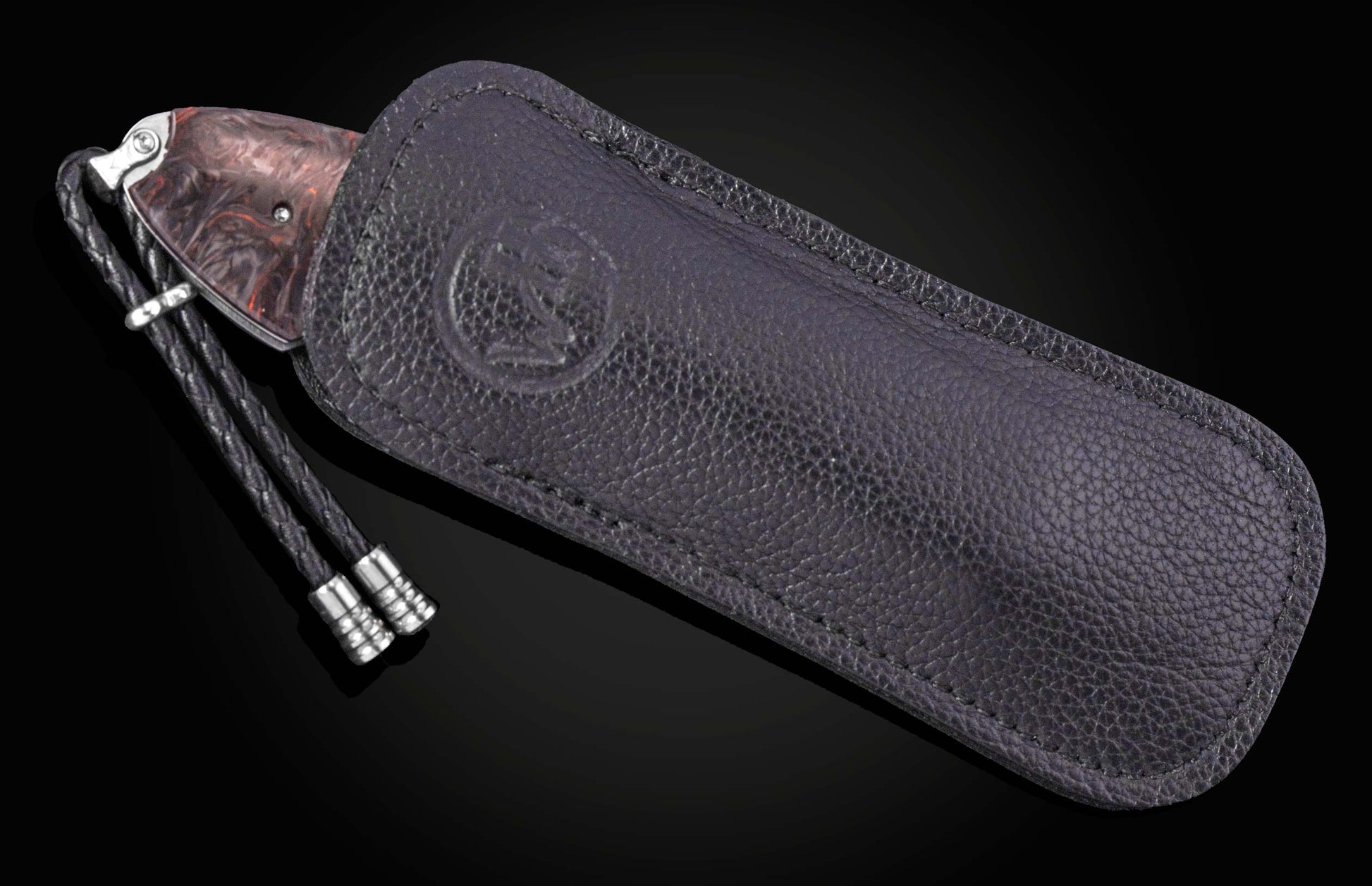 Red marbled handle knife with a black braided lanyard, partially inserted into a dark textured leather sheath with an embossed logo.