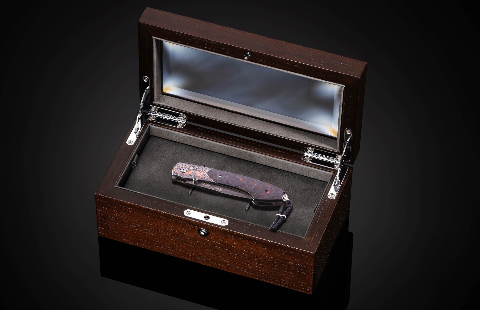 An intricate folding knife with a patterned handle and dark blade, presented in an open dark wood display box with a glass lid.