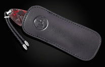 Red and black marbled handle knife, with a braided leather lanyard and silver tips, partially tucked into a black leather sheath with an embossed circular logo.