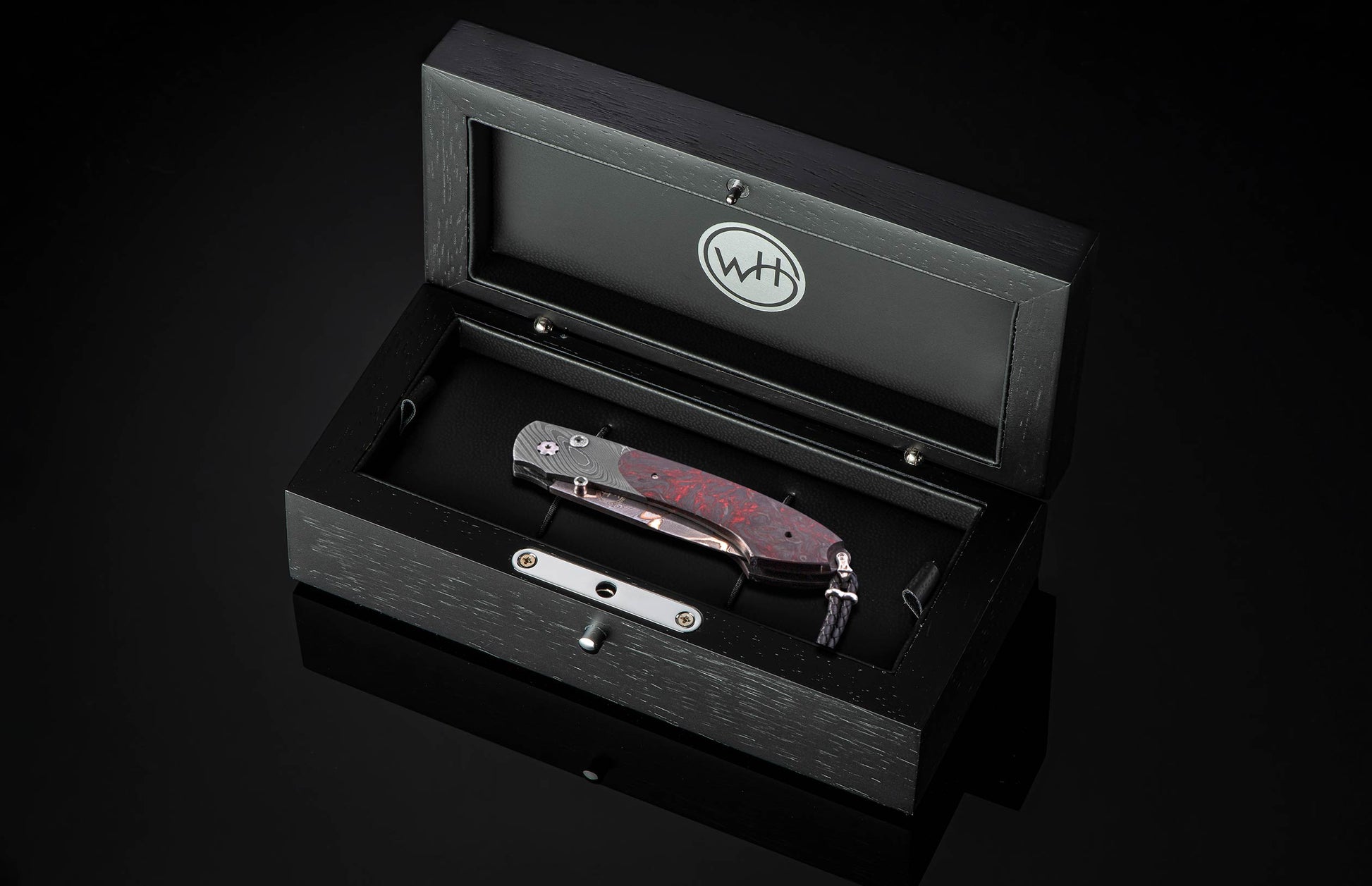 Folding knife with a red-patterned handle and patterned steel blade, displayed in an open black wooden box with a 'WH' logo.