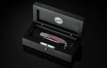 Folding knife with a red-patterned handle and patterned steel blade, displayed in an open black wooden box with a 'WH' logo.