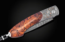 Folding knife with damascus blade and burl wood handle, featuring engraved quail design on silver bolster, side view on black background