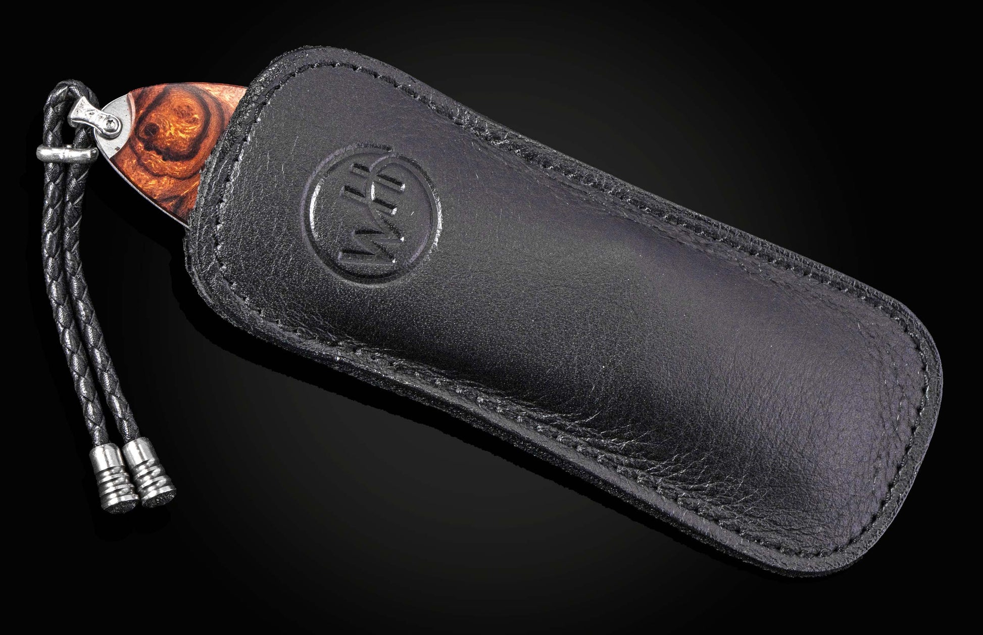 Handcrafted knife with warm wood handle partially sheathed in black leather, featuring embossed logo and braided cord, side view.