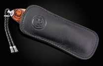 Handcrafted knife with warm wood handle partially sheathed in black leather, featuring embossed logo and braided cord, side view.