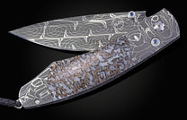 Partially open custom folder knife featuring a patterned Damascus blade, fossilized handle inlay, and blue gemstone accents.