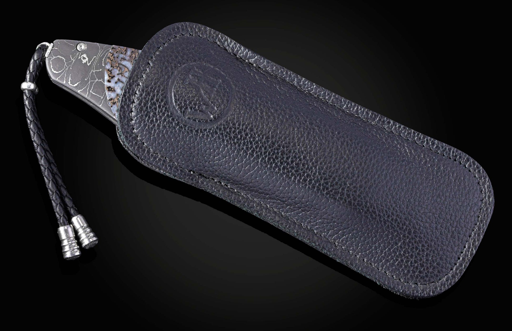B12 Venerable knife with patterned blade and stone inlay, partially in a dark pebbled leather sheath featuring an embossed logo and braided lanyard.