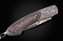 Western-style folding knife with damascus steel blade and handle, featuring intricate engraved bull and compass design, copper accents, and braided lanyard, side view.