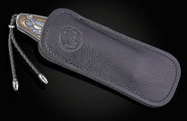 Ornate knife with a watch-face handle and braided leather lanyard, partially in a dark leather sheath with a debossed emblem.
