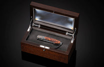 Luxury folding knife with burl wood handle and damascus blade in dark wood display case with glass lid, top-down view