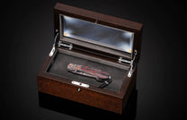 Luxury folding knife with vibrant red and black wood grain handle, displayed in polished dark wood presentation box with glass lid and leather-lined interior.