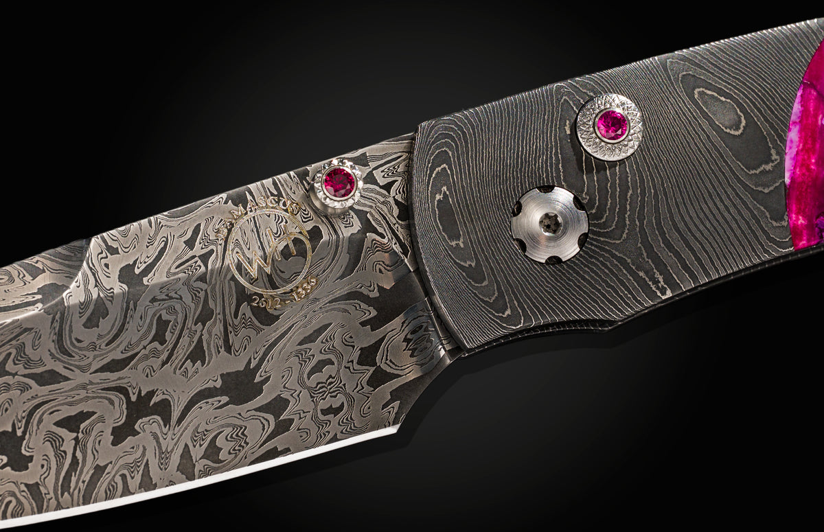 Damascus steel folding knife with ruby inlays and swirling pattern, close-up on black background.