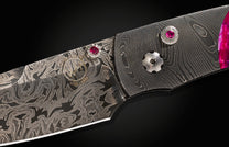 Damascus steel folding knife with ruby inlays and swirling pattern, close-up on black background.