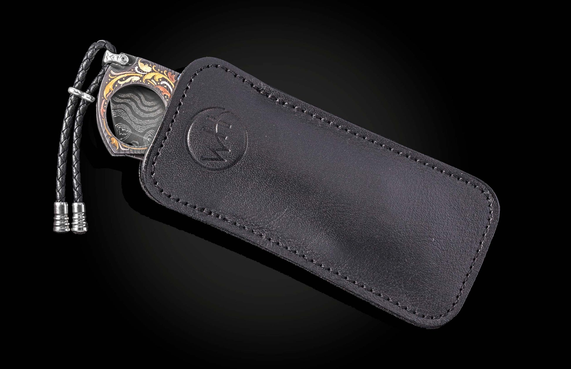Black leather cigar cutter with intricate gold and silver floral engravings, by NCT, featuring braided cord and embossed logo, side view.