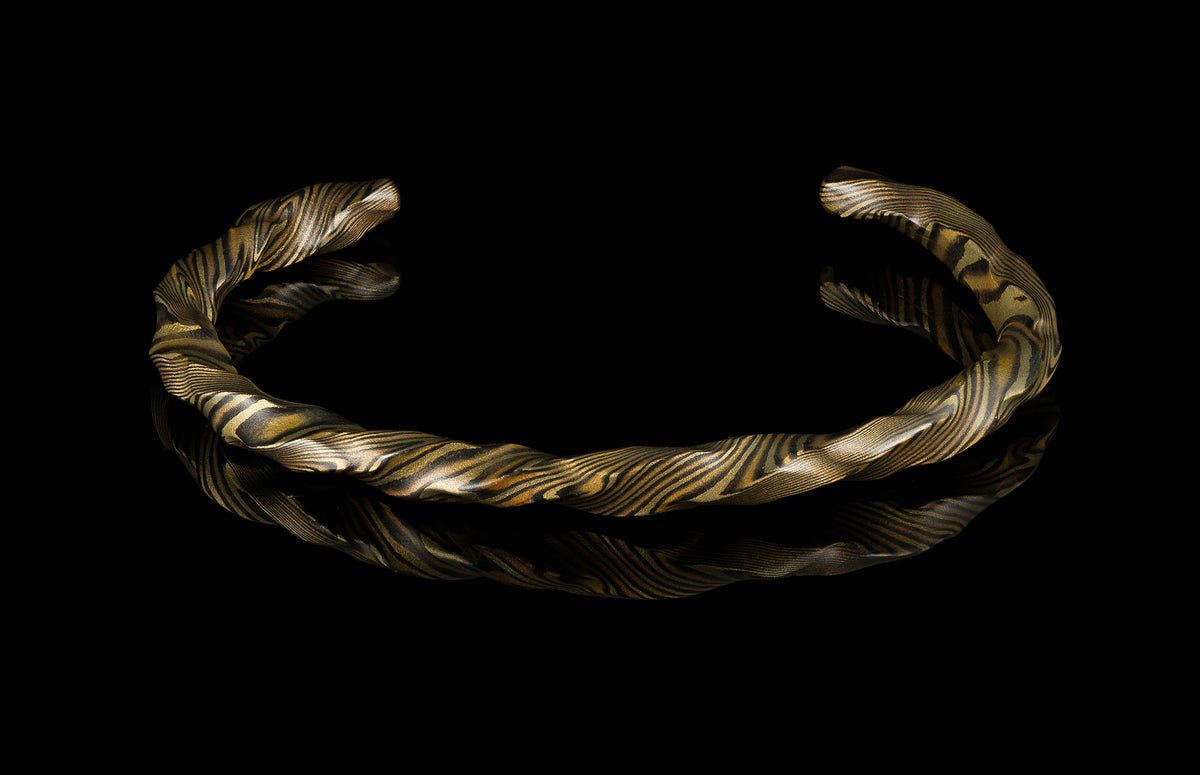 Twisted gold and black mokume-gane cuff bracelet by HC12 STRATA, showcasing intricate layered patterns and polished texture on reflective surface.