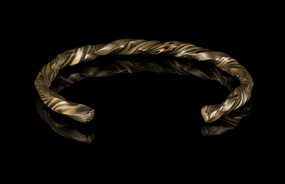 Twisted gold and black mokume-gane cuff bracelet by HC12, featuring layered metallic texture and reflective surface on dark background