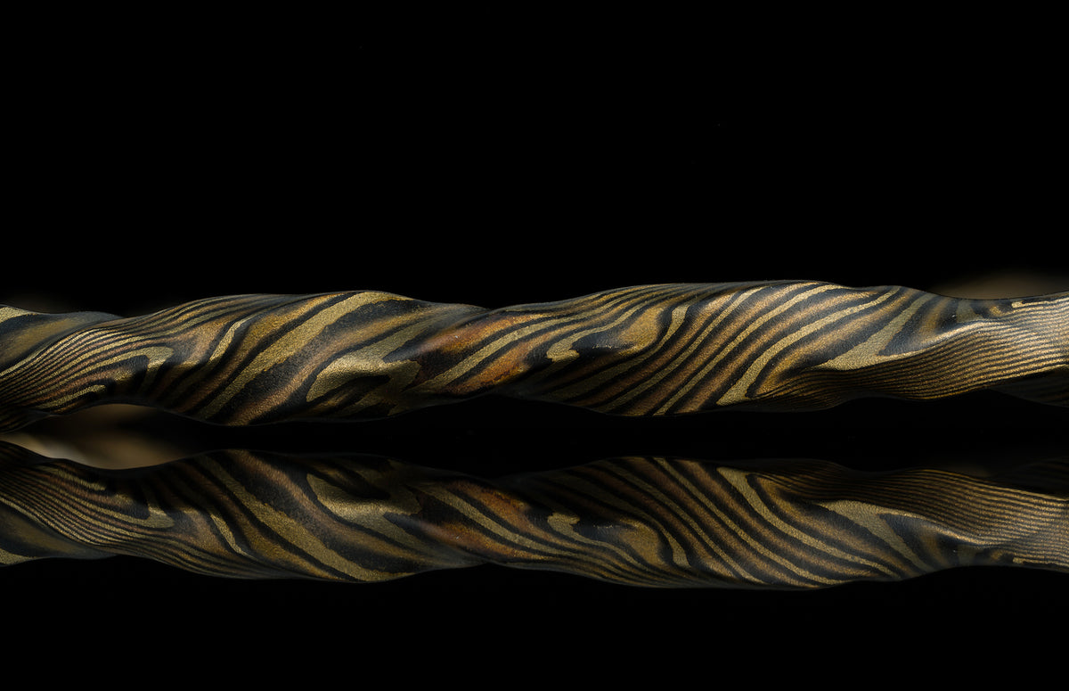 Twisted gold and black mokume-gane ring with layered wood-grain texture, reflective surface, side view on dark background