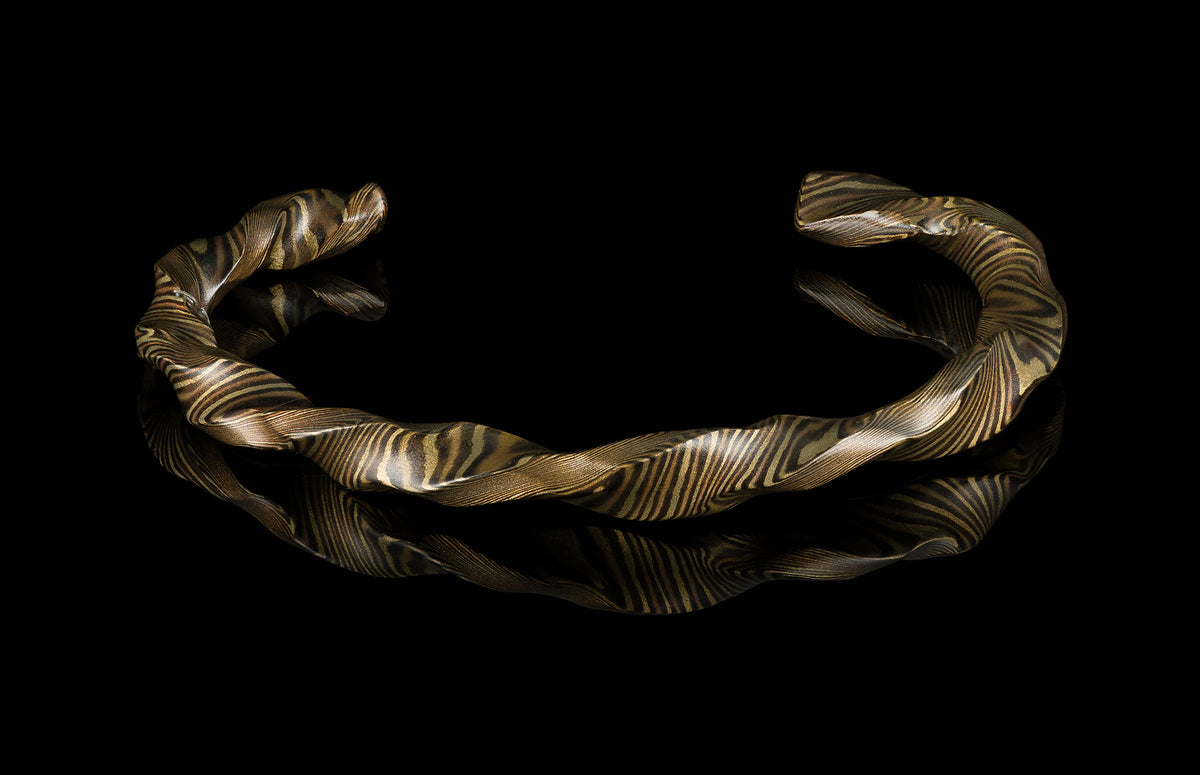 Twisted mokume-gane cuff bracelet by HC13 RIFTBORN, featuring golden and dark layered patterns, displayed on reflective black surface.