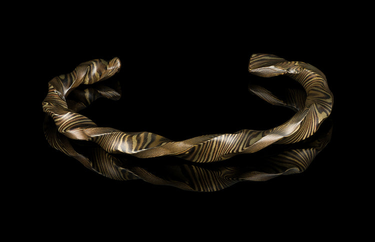 Twisted mokume-gane cuff bracelet by HC13 RIFTBORN, featuring golden and dark layered patterns, displayed on reflective black surface.