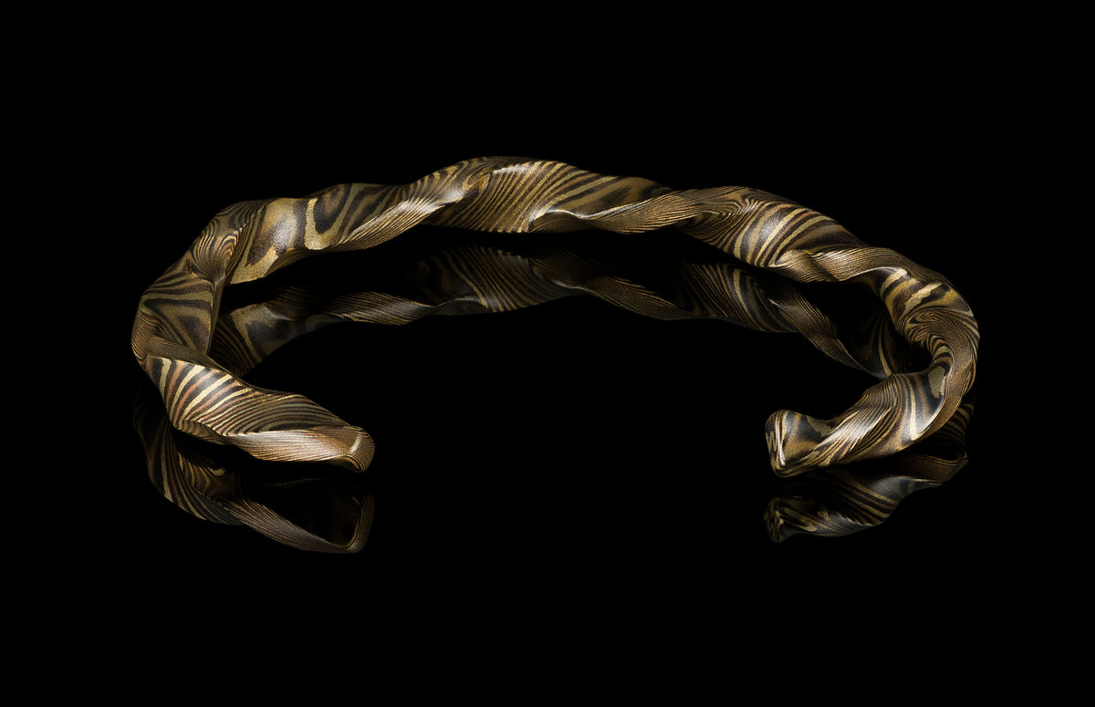 Twisted mokume-gane cuff bracelet by HC13 RIFTBORN, featuring golden and dark layered patterns, displayed on reflective black surface.