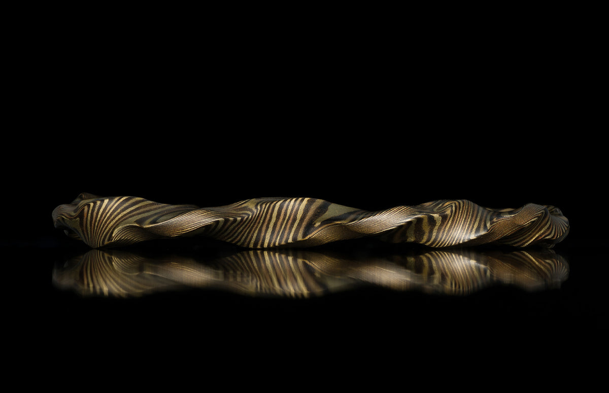 Twisted gold and dark metal mokume-gane ring with flowing striped pattern, side view on reflective black surface