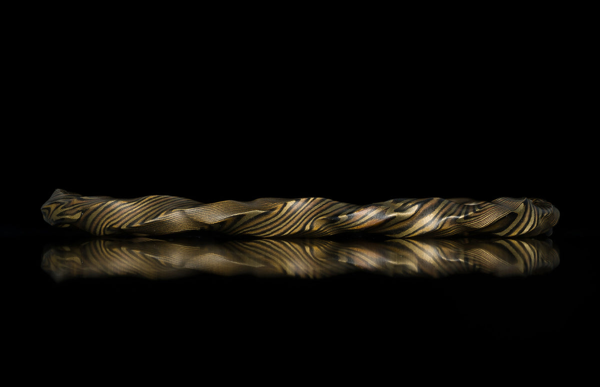 Twisted gold and dark patterned bracelet by HC13 RIFTBORN, showcasing intricate marbled texture with reflective surface, side view on black background