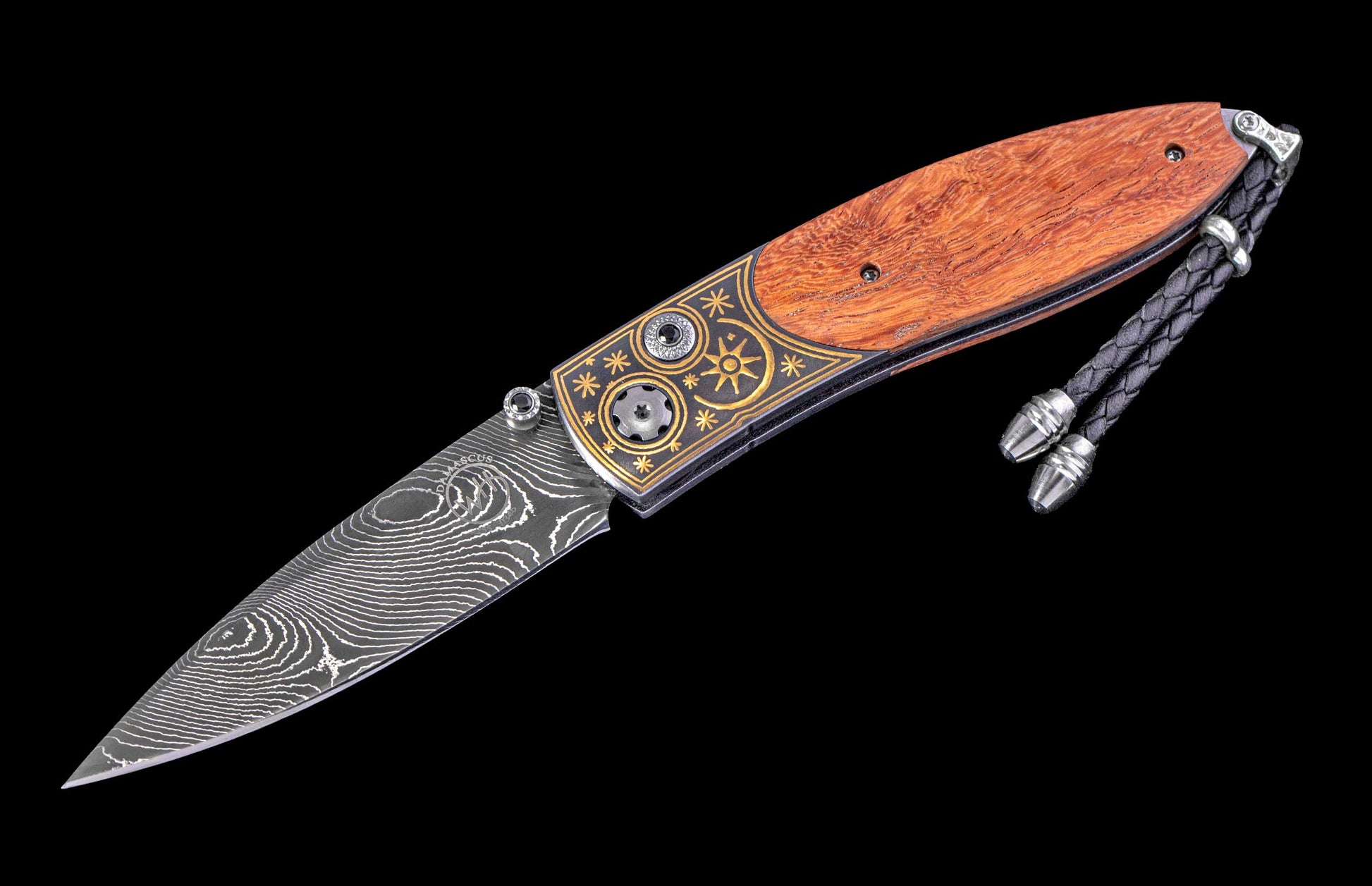 An open folding knife with a Damascus blade, rich wood handle, and ornate gold inlay on the black metal bolster, featuring a braided lanyard.