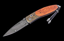 An open folding knife with a Damascus blade, rich wood handle, and ornate gold inlay on the black metal bolster, featuring a braided lanyard.
