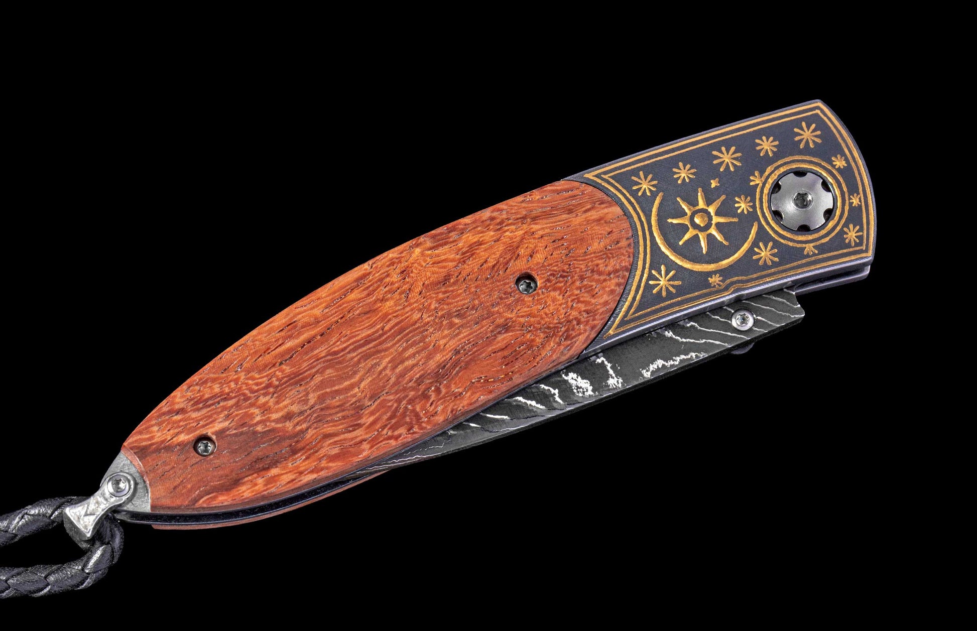 A closed folding knife with a reddish-brown wood handle, a dark gray bolster detailed with gold sun and star patterns, a Damascus blade, and a braided black leather lanyard.