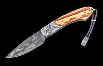 Folding knife with patterned Damascus steel blade, ornate engraved silver, light wood handle, blue gems, and a braided leather lanyard with wolf head bead.
