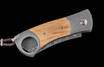 Closed folding knife with light wood and grey metal handle, gold pivot accent, Damascus steel blade, and braided leather lanyard.