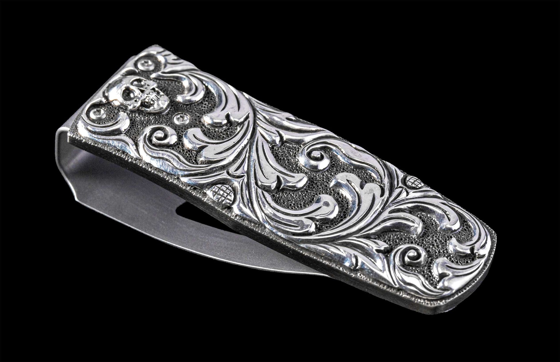 Silver metal money clip with an engraved skull, intricate scrollwork, and textured background, viewed at an angle.