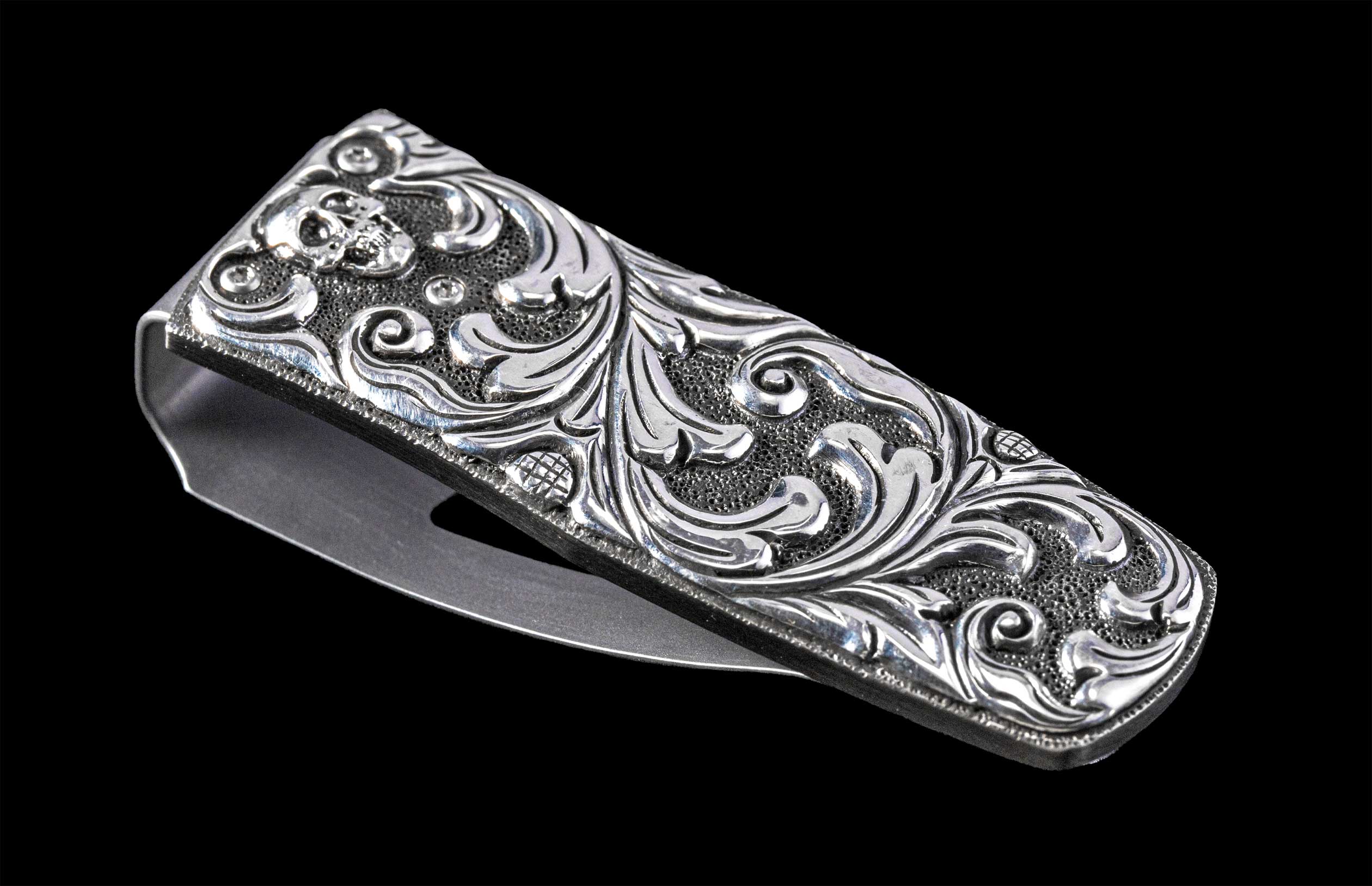 Silver metal money clip with an engraved skull, intricate scrollwork, and textured background, viewed at an angle.