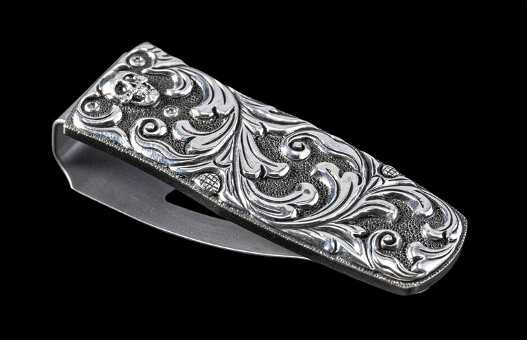 Silver metal money clip with an engraved skull, intricate scrollwork, and textured background, viewed at an angle.
