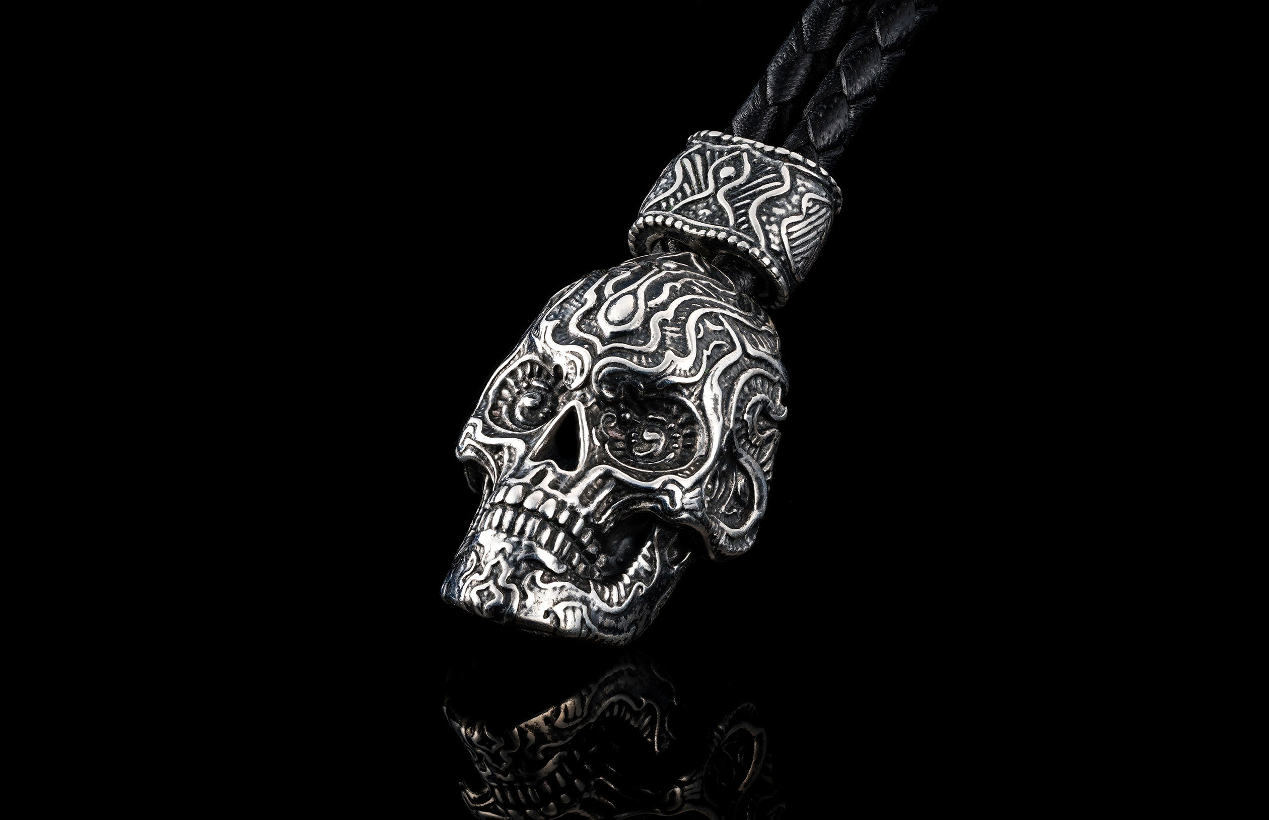 Silver metal skull key chain by TOUCHSTONE with intricate carvings, braided black leather cord, reflected on black surface.