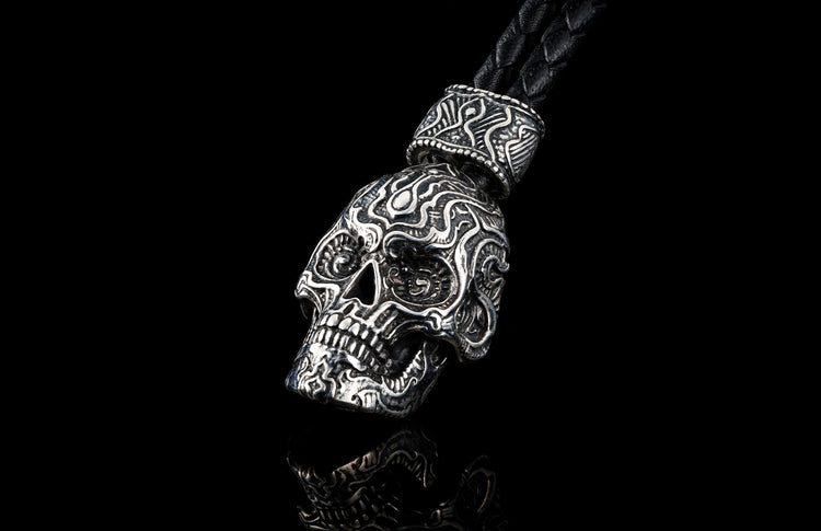 Silver metal skull key chain by TOUCHSTONE with intricate carvings, braided black leather cord, reflected on black surface.
